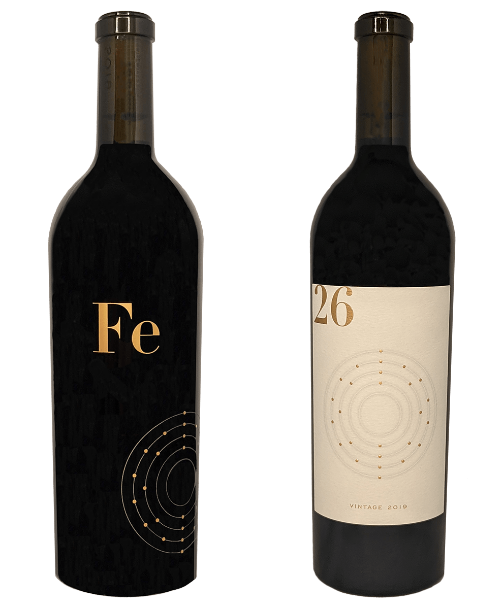 Two wine bottles, one with a black label featuring the symbol “Fe” and one with a cream label featuring the number “26,” both displaying a diagram of an iron atom.