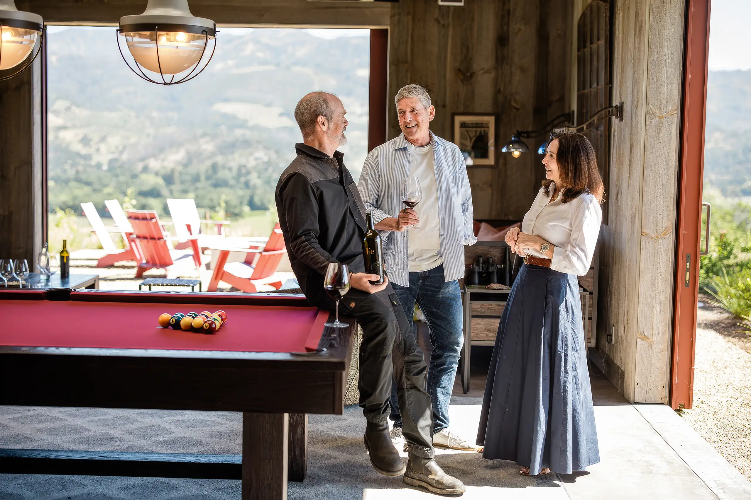 Aaron, Jon, and Joanne talk while standing around a pool table in a room with a large open doorway overlooking a hillside.