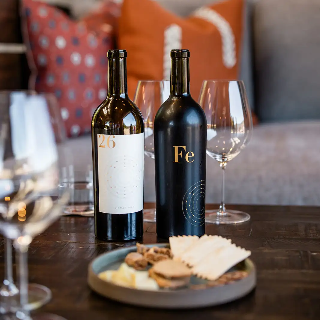 Two wine bottles, one with a white label reading “26” and one black bottle labeled “Fe,” sitting on a table with wine glasses and a small plate of crackers and cheese.